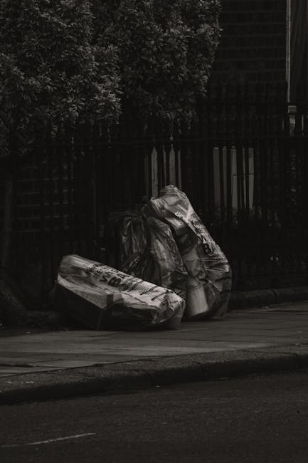 Three large black plastic rubbish bags filled with waste are placed on the pavement next to a black metal fence, with a tall hedge visible behind it. The bags appear to contain household or garden ref