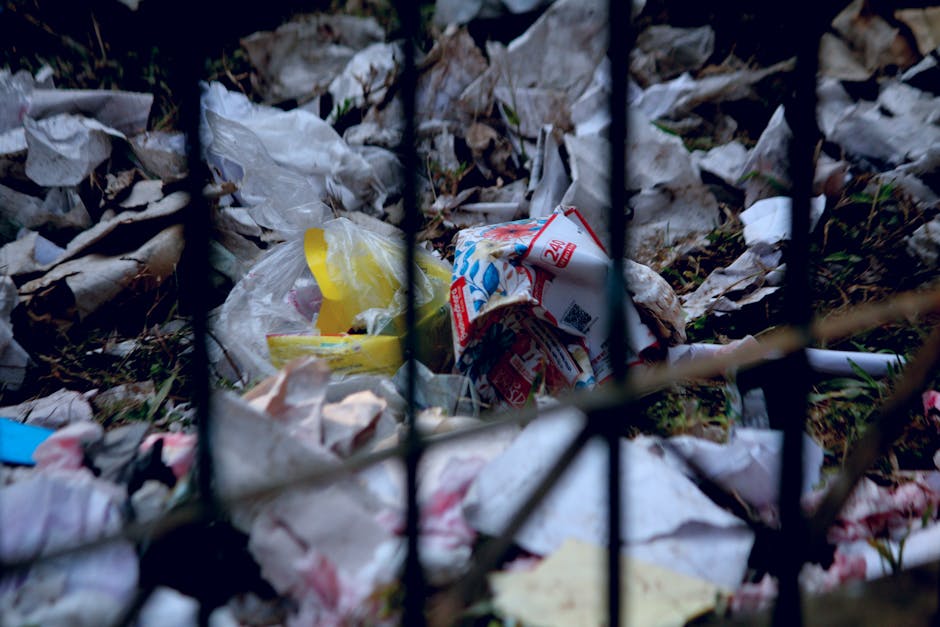 A close-up view of a metal fence with vertical black bars, behind which lies a pile of scattered rubbish consisting of crumpled paper, plastic wrappers, and plastic bags in various colors, including w