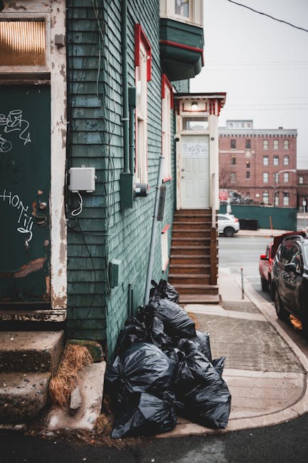 A close-up view of a metal fence with vertical black bars, behind which lies a pile of scattered rubbish consisting of crumpled paper, plastic wrappers, and plastic bags in various colors, including w